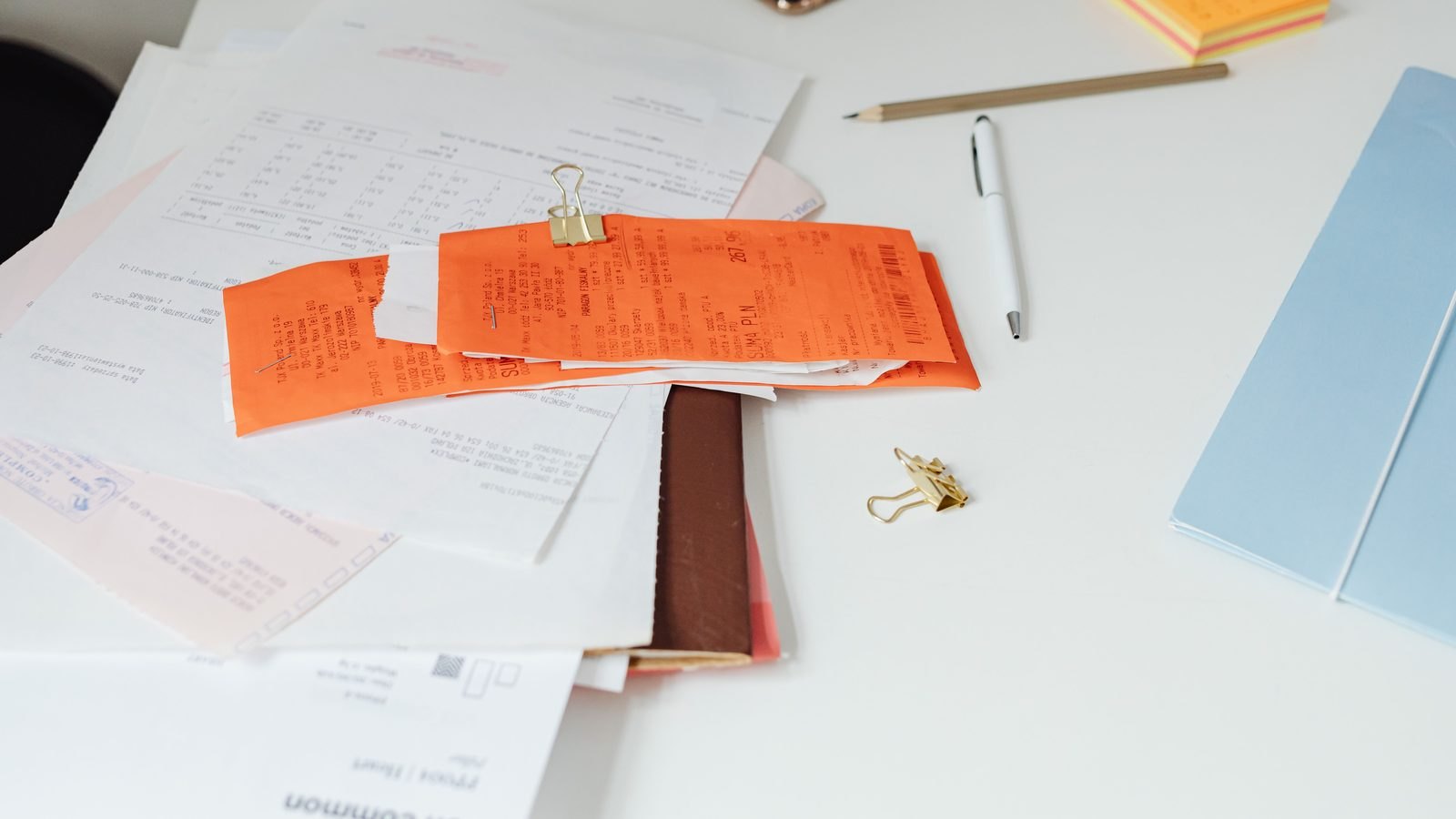 Mesa branca coberta de papelada de pequeno negócio: notas fiscais laranja presas por clipe de metal dourado, comprovantes e formulários brancos espalhados embaixo, uma caneta e um lápis de lado, um bloco de papel azul e um bloco de recados colorido no canto.