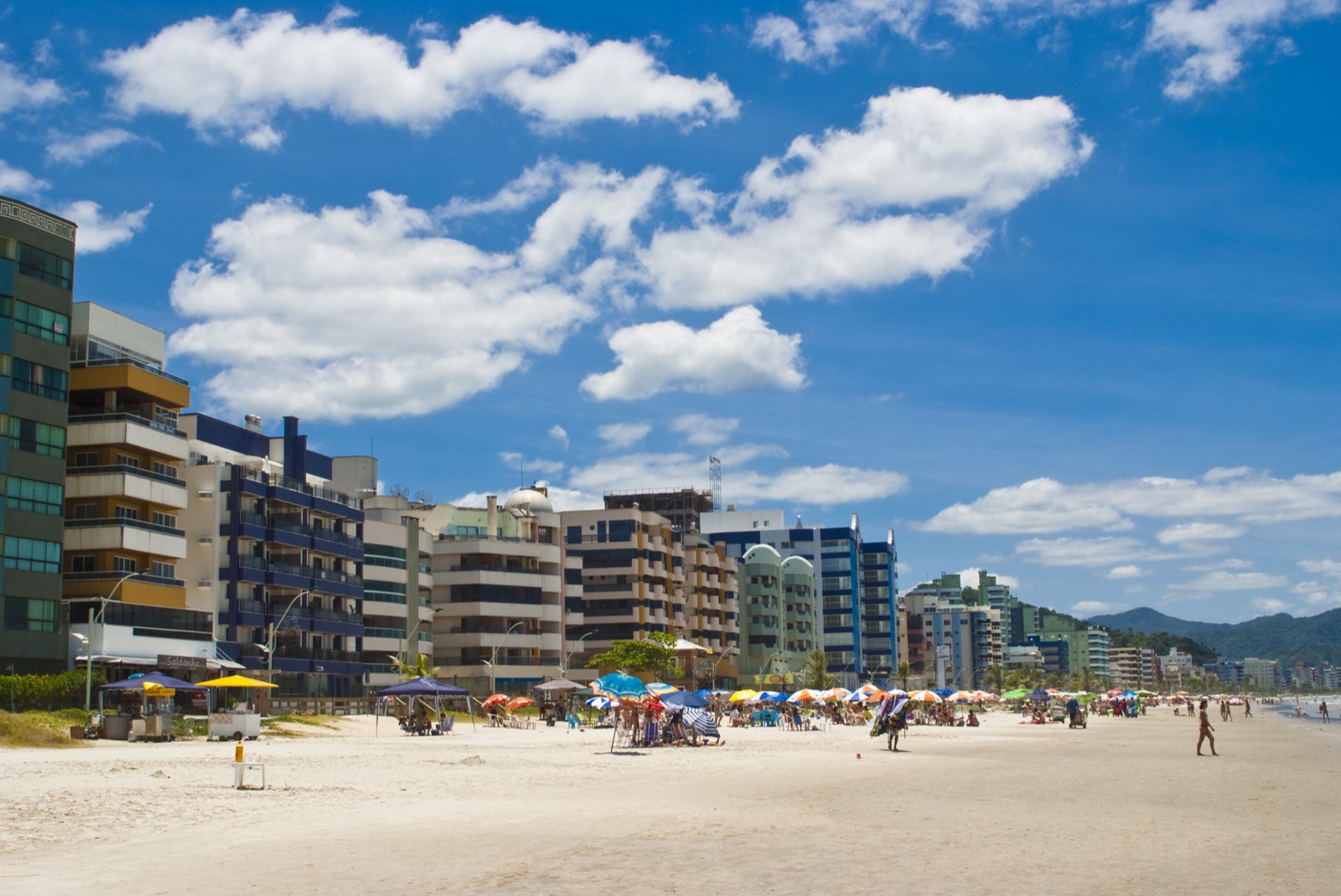 Faixa de areia da Meia Praia vista do nível da praia, com guarda-sóis coloridos e, à esquerda, a linha contínua de prédios residenciais de vários andares que ocupa toda a orla.