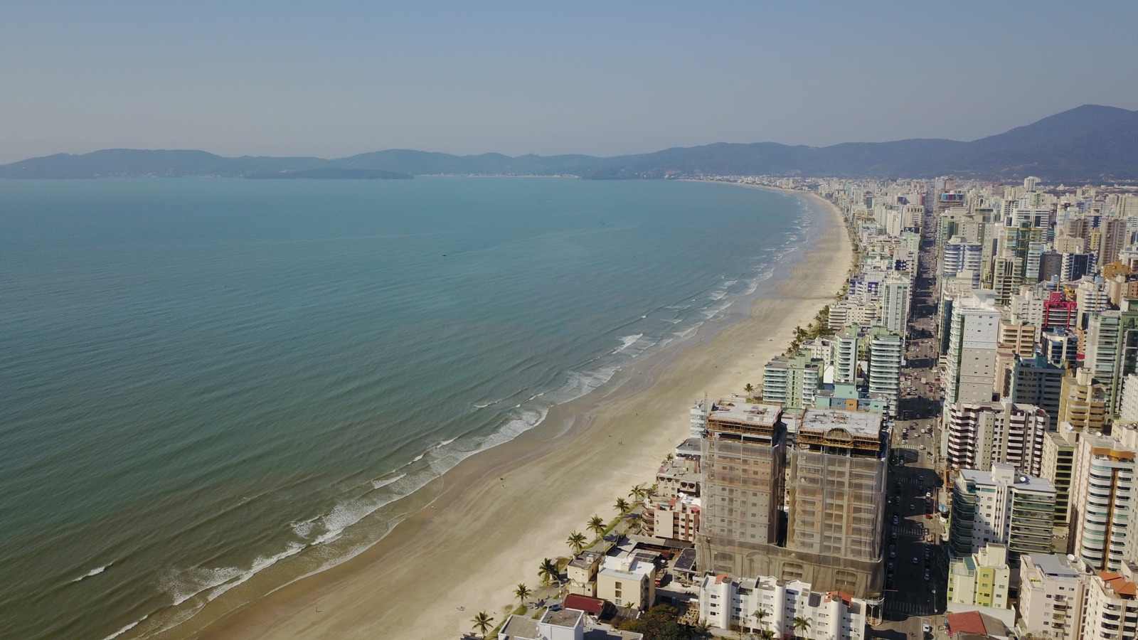 Vista aérea da Meia Praia em Itapema: o mar ocupa a metade esquerda da imagem, a faixa de areia curva acompanha a orla e, à direita, a linha contínua de edifícios altos se estende até os morros ao fundo.