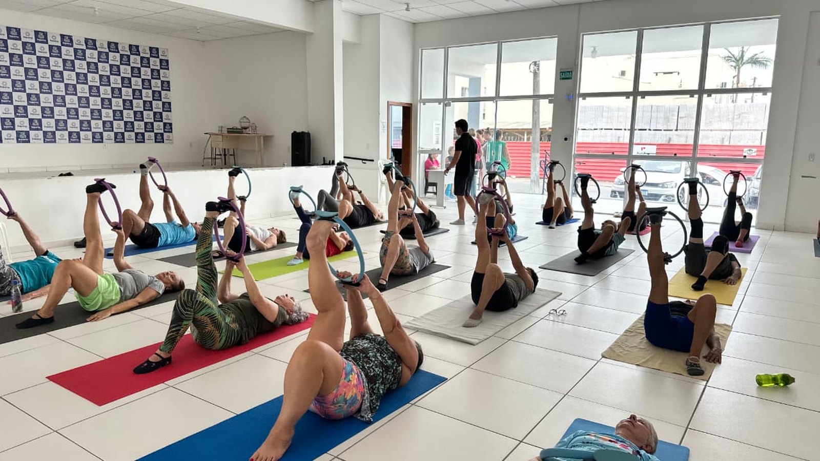 Turma de pilates para pessoas idosas numa sala ampla e clara, com participantes deitados em colchonetes coloridos segurando arcos de exercício com os pés levantados.