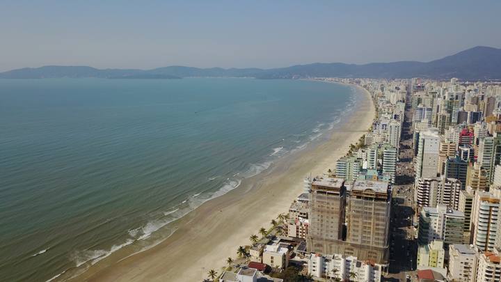 Vista aérea da Meia Praia em Itapema: o mar ocupa a metade esquerda da imagem, a faixa de areia curva acompanha a orla e, à direita, a linha contínua de edifícios altos se estende até os morros ao fundo.