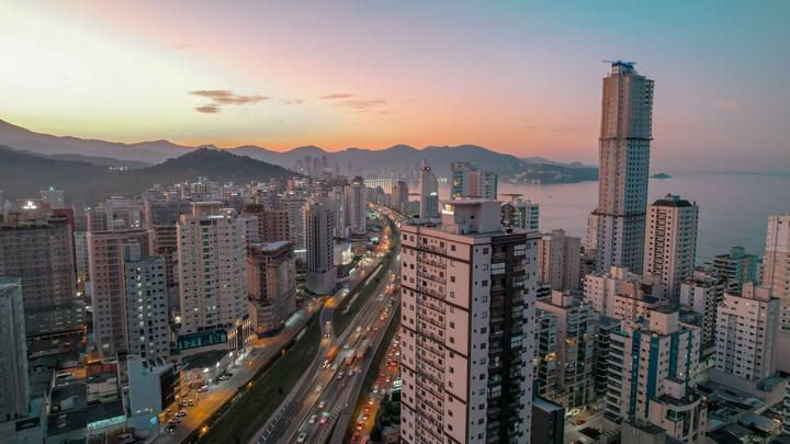 Itapema vista do alto ao pôr do sol: torres residenciais densas ocupam toda a imagem, uma delas ainda em obra com guindaste no topo, a rodovia corta o meio e o mar aparece à direita, com morros no horizonte.