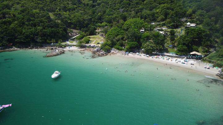 Vista aérea de uma enseada de Porto Belo: água esverdeada e transparente ocupa a metade inferior da imagem, uma lancha branca está ancorada e, na faixa de areia ao fundo, guarda-sóis se espalham diante de um morro coberto de mata fechada.