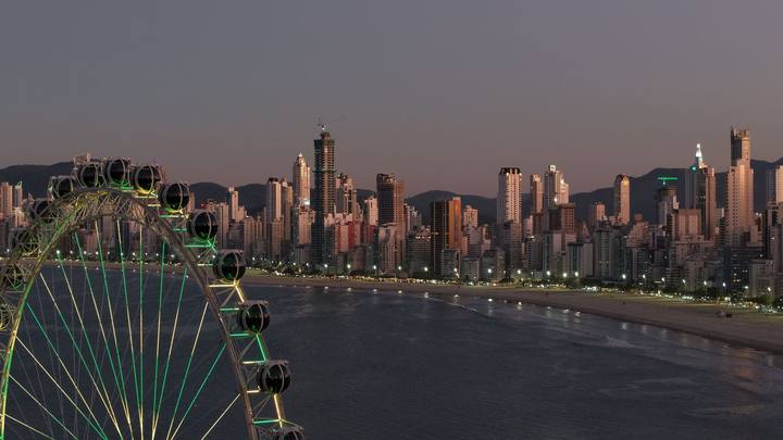 Balneário Camboriú ao anoitecer vista do alto: a faixa de prédios altos acompanha a curva da praia, com uma roda-gigante iluminada em primeiro plano e o mar escuro à direita.