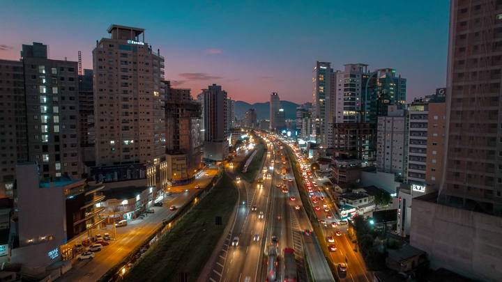 Itapema ao anoitecer vista do alto: a BR-101 corta a imagem ao meio com as duas pistas cheias de carros de faróis acesos, e prédios altos se erguem dos dois lados.