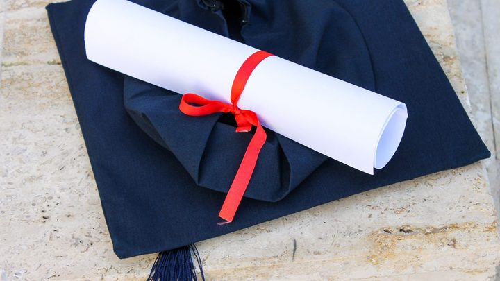 Capelo de formatura azul-escuro apoiado sobre uma superfície de pedra clara, com um diploma enrolado amarrado por uma fita vermelha em cima dele.