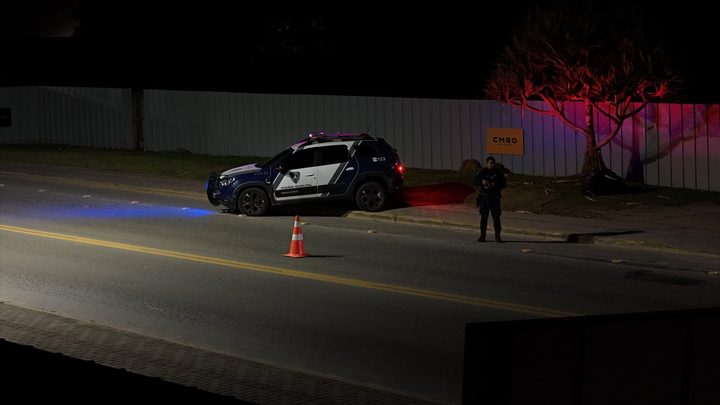 Viatura da Guarda Municipal de Porto Belo parada à beira da rua à noite, com giroflex azul e vermelho aceso, um cone laranja no asfalto e um agente fardado em pé na calçada ao lado.