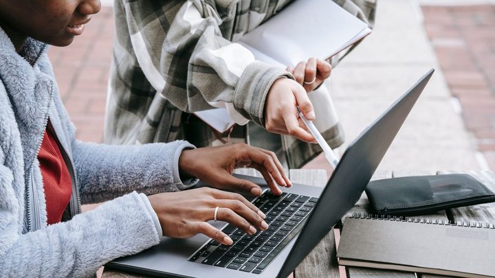 Duas estudantes sentadas num banco de praça digitando num notebook aberto sobre a mesa, com cadernos empilhados ao lado e uma delas apontando a tela com a caneta.