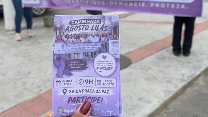 Mão segurando o panfleto lilás da Caminhada do Agosto Lilás, com a data de 27 de agosto, o horário das 9h e a saída da Praça da Paz impressos, e a faixa da campanha desfocada ao fundo, na calçada.