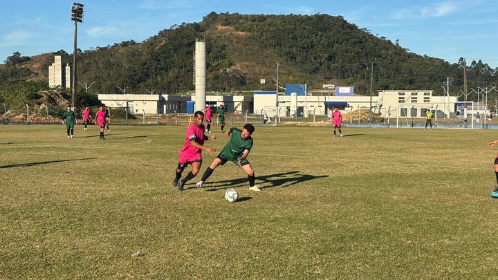 Dois jogadores de futebol amador disputam a bola no meio do campo gramado, um de uniforme rosa e outro de verde, com outros atletas espalhados ao fundo e um morro coberto de mata atrás do alambrado.