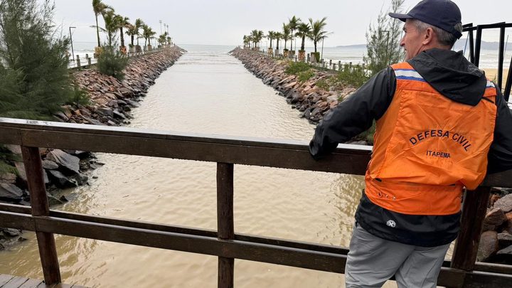 Agente da Defesa Civil de Itapema, de colete laranja e boné, apoiado no corrimão de uma passarela de madeira molhada, olhando a água barrenta que corre entre dois molhes de pedra até o mar, em dia nublado.