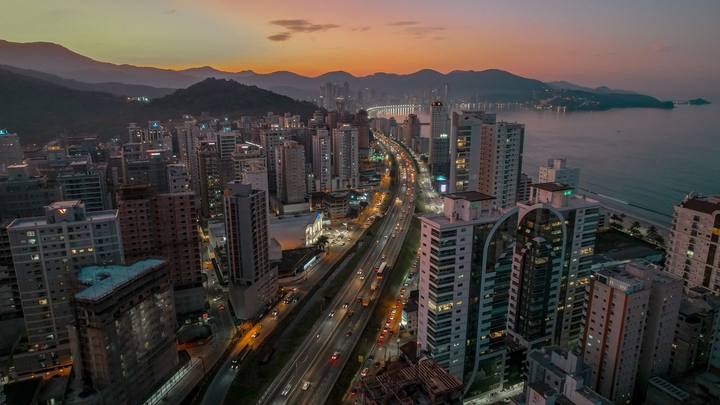 Itapema vista do alto no fim da tarde, com as luzes da cidade começando a acender ao longo da orla e o mar ocupando o lado esquerdo da imagem.