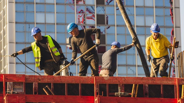 Quatro operários de capacete azul trabalhando sobre a laje de um prédio em obra, um puxando um cabo e outro segurando a mangueira de concreto, com a fachada espelhada de um edifício ao fundo.