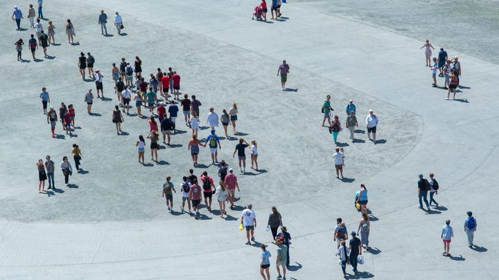 Vista de cima de uma praça de piso claro em dia de sol, com dezenas de pessoas caminhando espalhadas pelo chão, algumas em grupo e outras sozinhas, projetando sombras curtas.