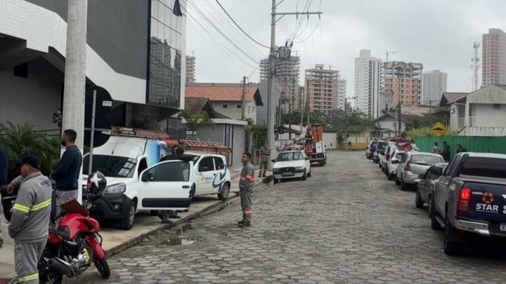 Rua de paralelepípedo em Porto Belo num dia nublado, com poste de energia carregando um feixe de cabos que cruza a via, carros estacionados dos dois lados e trabalhadores de uniforme cinza parados na calçada.