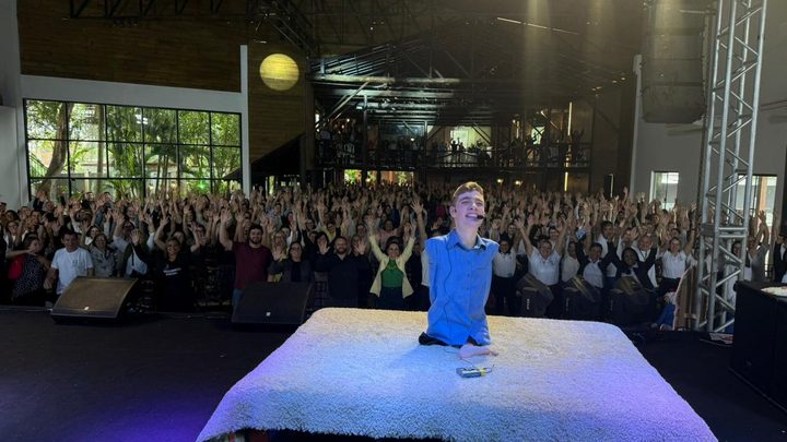 Palestrante no centro de um palco, apoiado sobre um praticável forrado de tapete claro, de camisa azul e microfone de haste, sorrindo diante de uma plateia de professores que aplaude com os braços erguidos.