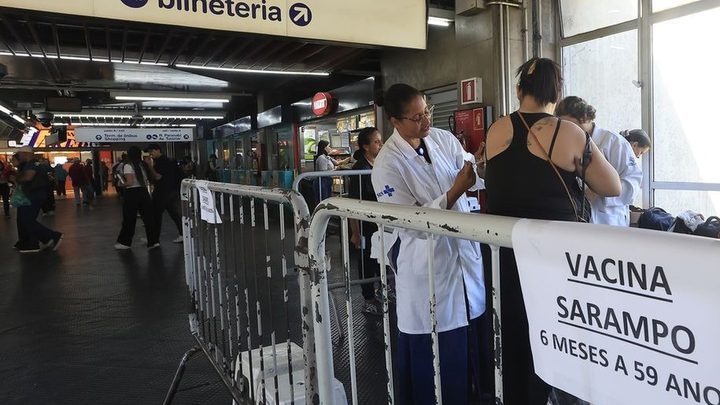 Posto de vacinação montado no saguão de uma estação de metrô, com profissionais de jaleco branco atrás de grades metálicas e uma faixa branca com os dizeres vacina sarampo, 6 meses a 59 anos.