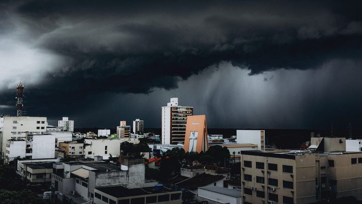 Nuvem de temporal muito escura e baixa cobrindo o céu inteiro sobre os prédios de uma cidade, com uma cortina de chuva descendo ao fundo à direita e uma torre de antena recortada contra o cinza à esquerda.
