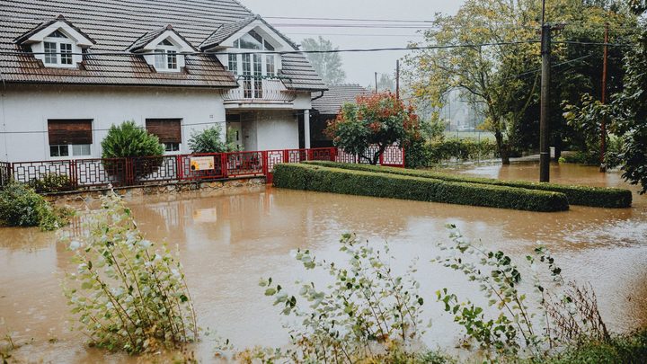 Casa de telhado escuro com a frente tomada por água barrenta que cobre o jardim, o portão vermelho e parte da rua, sob chuva forte, com poste de energia e árvores ao fundo.