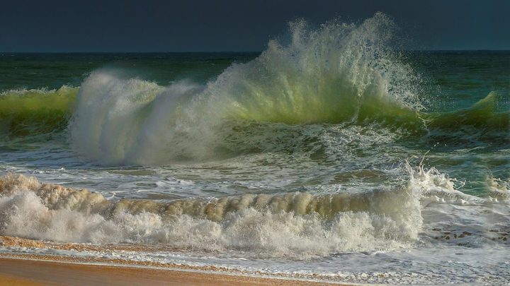 Onda alta quebrando perto da beira de uma praia de areia, com a crista branca levantada pelo vento e o céu escuro carregado ao fundo, sem nenhuma pessoa na água.