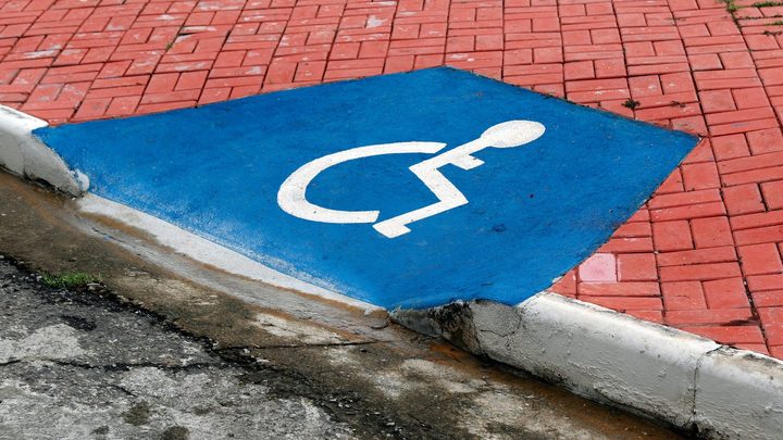 Rebaixamento de meio-fio pintado de azul com o símbolo branco de acessibilidade, ligando a calçada de piso vermelho ao asfalto de uma rua, sem nenhuma pessoa no quadro.