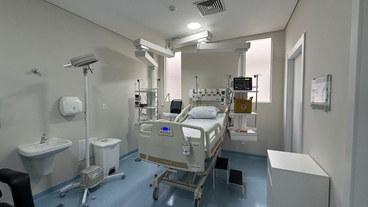 Box individual da nova UTI do Hospital Santo Antônio de Itapema, com leito hospitalar de grades brancas, monitor multiparamétrico ligado na parede, braços articulados presos ao teto, pia e lixeiras, sem ninguém no quadro.