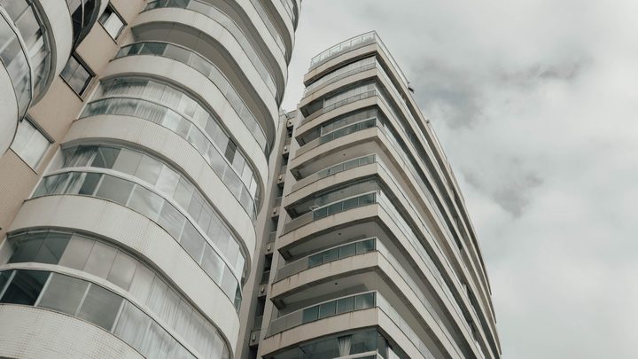 Fachada de dois prédios residenciais vistos de baixo para cima, com sacadas arredondadas fechadas com vidro e telas de proteção, sob um céu nublado.