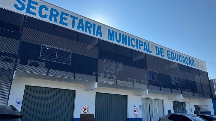 Fachada da Secretaria Municipal de Educação de Itapema, prédio de dois andares com o nome em letras azuis no alto, grades escuras no pavimento superior e portões verdes na frente.