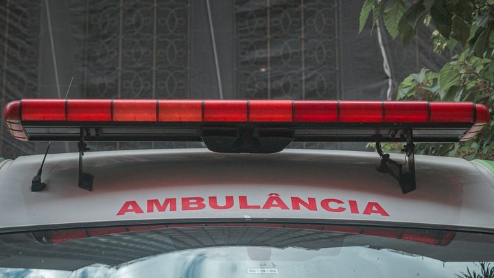 Vista frontal do teto de uma ambulância branca, com a barra de sirene vermelha atravessando a imagem e a palavra AMBULÂNCIA escrita em letras vermelhas sobre o capô.