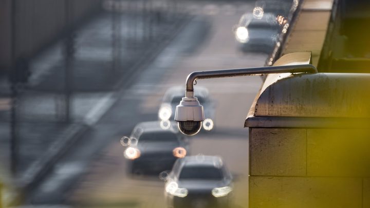 Câmera de segurança em formato de domo presa a um braço de metal sobre uma via urbana, com carros de faróis acesos passando desfocados ao fundo e uma mureta amarela em primeiro plano.