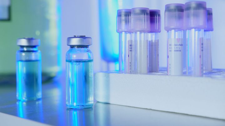 Dois frascos de vidro fechados com lacre de alumínio e cheios de líquido azul sobre uma bancada branca de laboratório, ao lado de uma grade de suporte com tubos de coleta de tampa roxa.
