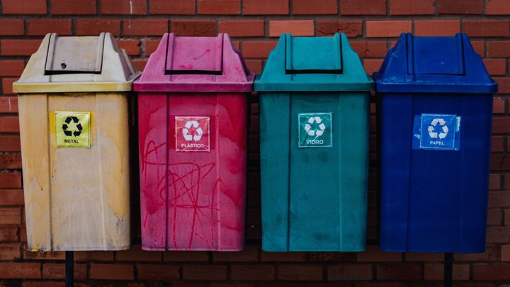 Quatro lixeiras de coleta seletiva enfileiradas diante de uma parede de tijolos, com adesivos do símbolo da reciclagem e as palavras metal, plástico, vidro e papel, nas cores bege, rosa, verde e azul.