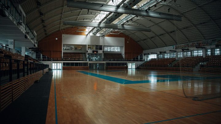 Ginásio poliesportivo vazio visto da lateral da quadra, com piso de madeira clara, linhas azuis pintadas no chão, arquibancadas de cadeiras laranjas dos dois lados e teto curvo de estrutura metálica.