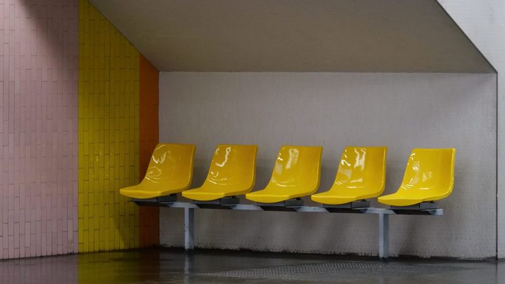 Fileira de cinco cadeiras amarelas de plástico presas a uma estrutura de metal, encostadas na parede clara de um corredor vazio de prédio público, sem ninguém sentado.