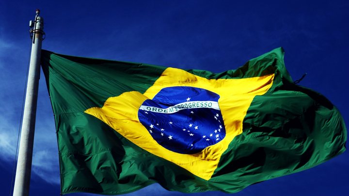 Bandeira do Brasil hasteada em mastro de metal, tremulando aberta pelo vento contra um céu azul sem nuvens.