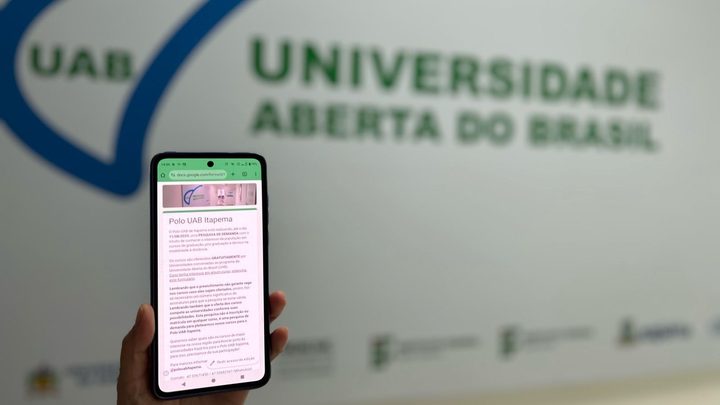 Uma mão segura um celular com o formulário do Polo UAB Itapema aberto na tela, diante da parede branca do polo com o logotipo da Universidade Aberta do Brasil em azul e verde.