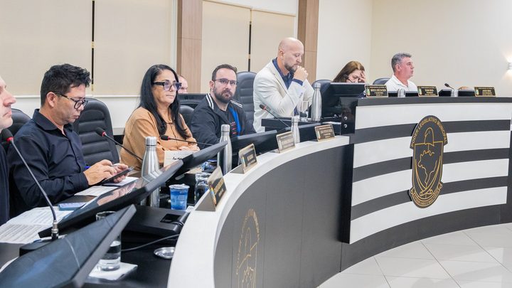 Vereadores sentados na bancada em curva do plenário da Câmara de Itapema durante sessão, com microfones, placas de identificação e o brasão do Poder Legislativo Municipal na lateral.