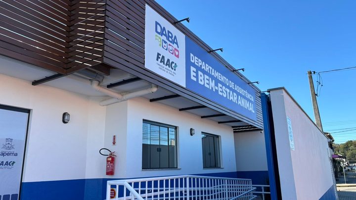 Fachada da sede do DABA em Itapema, pr&eacute;dio branco com faixa azul na parte de baixo e letreiro azul escrito Departamento de Assist&ecirc;ncia e Bem-Estar Animal sobre a entrada, com uma rampa de corrim&atilde;o branco na frente.