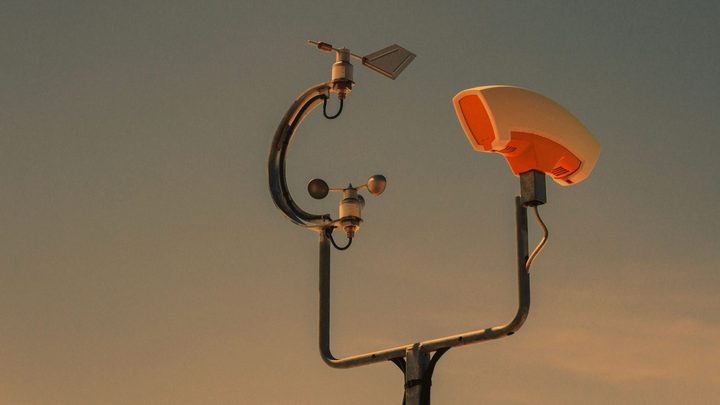 Topo de uma estação meteorológica recortada contra o céu no fim da tarde, com o cata-vento de direção e o anemômetro de três conchas presos a um braço curvo de metal e, ao lado, o abrigo branco do sensor de temperatura.