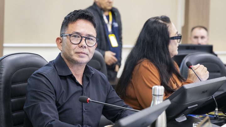 Vereadores sentados nas cadeiras do plenário da Câmara de Itapema durante sessão, com microfones de haste sobre a bancada, monitores de computador à frente e um servidor de crachá em pé ao fundo.