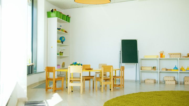 Sala de educação infantil vazia, com uma mesa baixa cercada de cadeirinhas de madeira, estante branca com brinquedos e tapete redondo verde no chão claro.