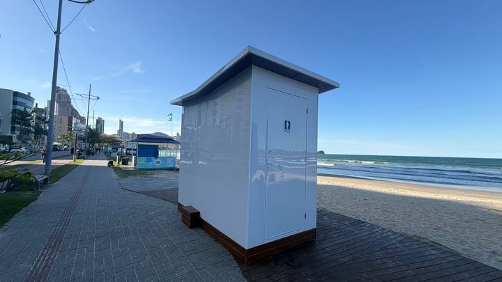 Módulo sanitário branco de porta única já instalado sobre o deque de madeira do calçadão da orla de Itapema, com a faixa de areia e o mar à direita e a linha de prédios da avenida à esquerda.