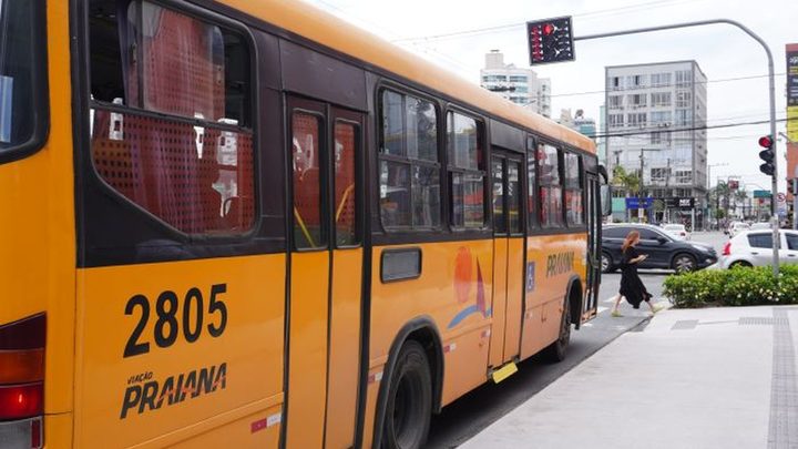 Ônibus amarelo da Viação Praiana, de número 2805, parado junto ao meio-fio de uma avenida de Itapema, com semáforo, prédios altos e comércio ao fundo.