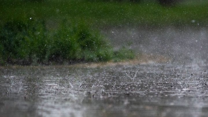 Chuva forte batendo no asfalto encharcado, com as gotas levantando respingos brancos na lâmina de água e uma faixa de vegetação verde desfocada ao fundo.