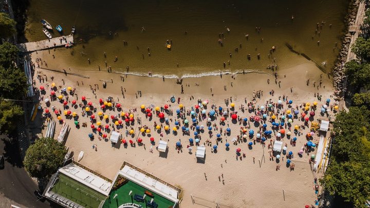 Vista de drone de uma faixa de praia cheia, com dezenas de guarda-sóis coloridos enfileirados sobre a areia clara, banhistas dentro da água rasa e um trapiche na ponta do quadro.