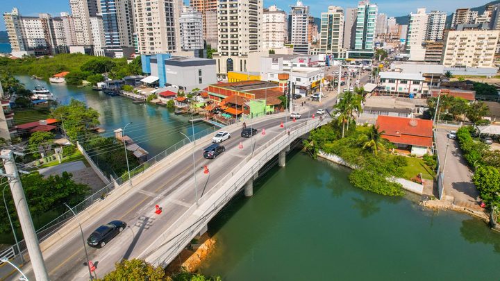 Vista aérea de Itapema com a ponte sobre o rio, carros passando entre cones laranja, casas e comércios de um lado e a linha de prédios altos ao fundo.