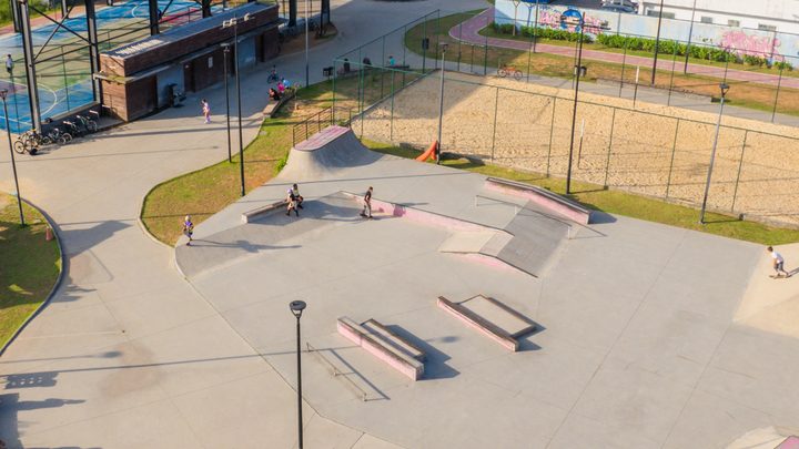 Vista aérea de área de lazer pública em Itapema, com pista de skate de concreto e rampas, quadra de areia cercada por alambrado ao lado e ginásio coberto ao fundo.