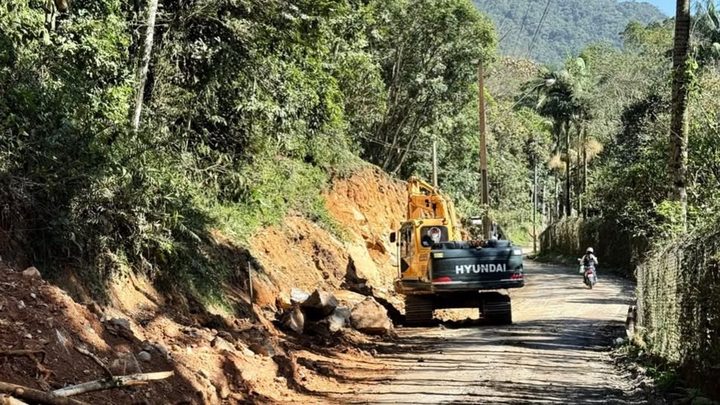 Escavadeira amarela cortando o barranco de terra vermelha na margem de uma estrada rural estreita e sem asfalto no Alto Areal, em Itapema, com blocos de rocha soltos no chão e mata fechada dos dois lados.