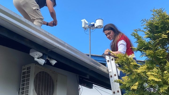 Dois técnicos instalam uma estação meteorológica no telhado de uma escola sob céu azul: um homem agachado sobre a calha aperta uma peça e uma mulher de colete vermelho trabalha no alto de uma escada de alumínio, com o mastro da estação e o pluviômetro branco ao fundo.