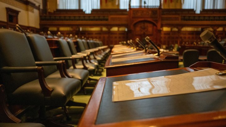 Plenário vazio de uma casa legislativa, com fila de poltronas de couro verde encostadas em bancadas de madeira escura, microfones de haste sobre cada lugar e papéis apoiados no tampo.